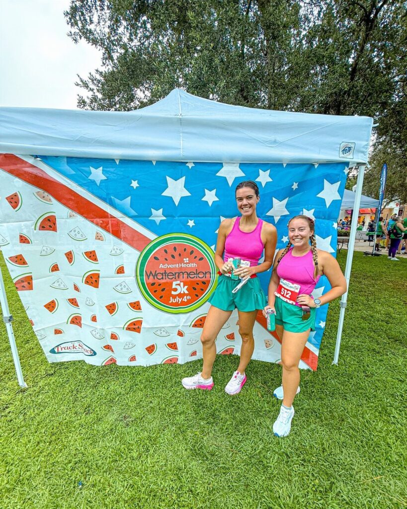 Two women in pink and green running outfits posing before a patriotic backdrop.