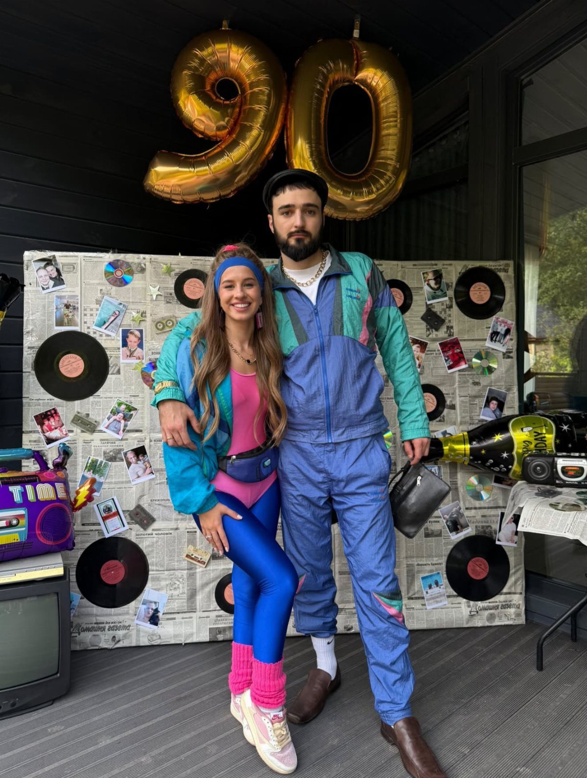 Couple posing in colorful 90s athletic outfits against a music-themed party backdrop.