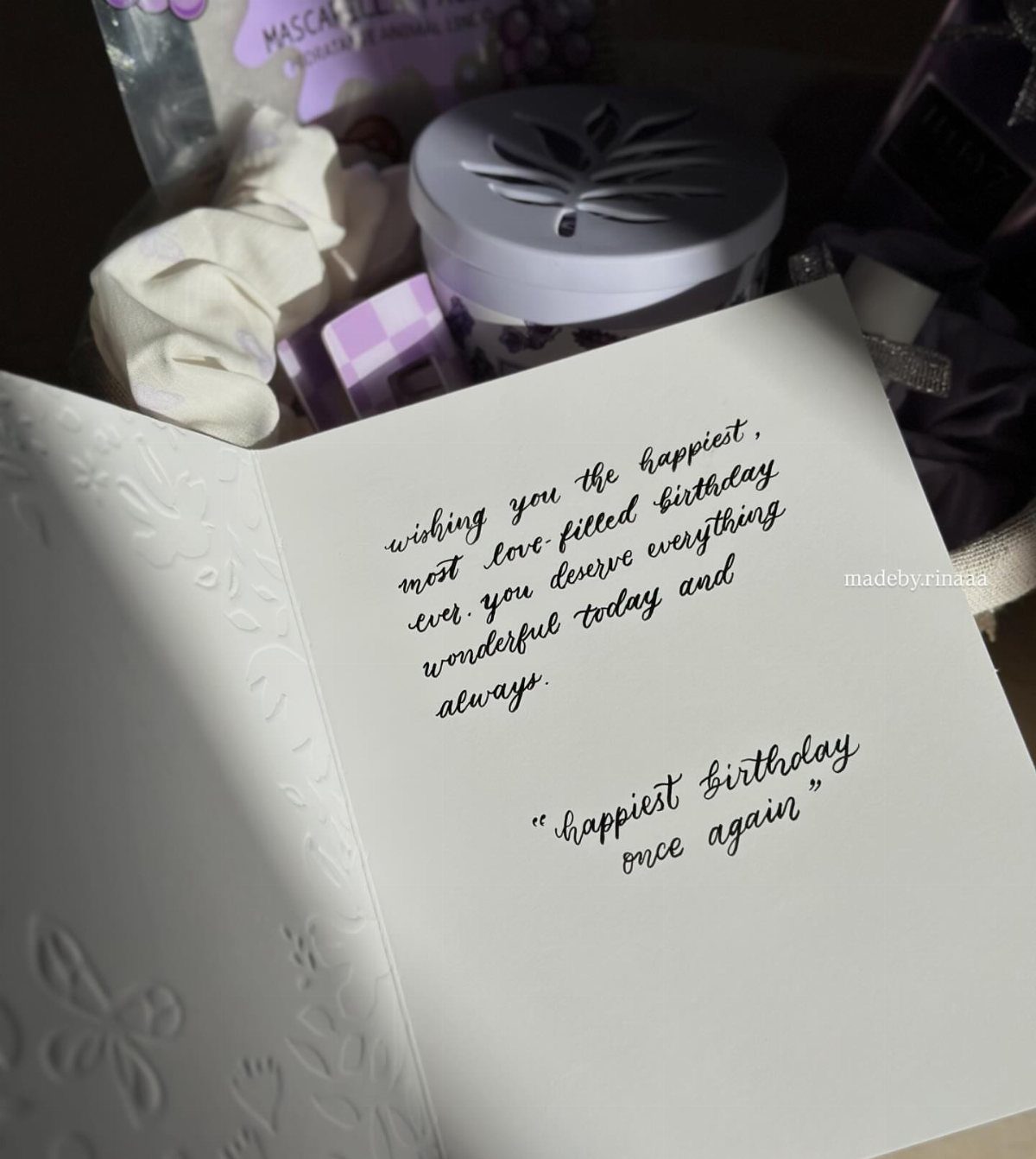 Open white greeting card with handwritten birthday message and gift items in background