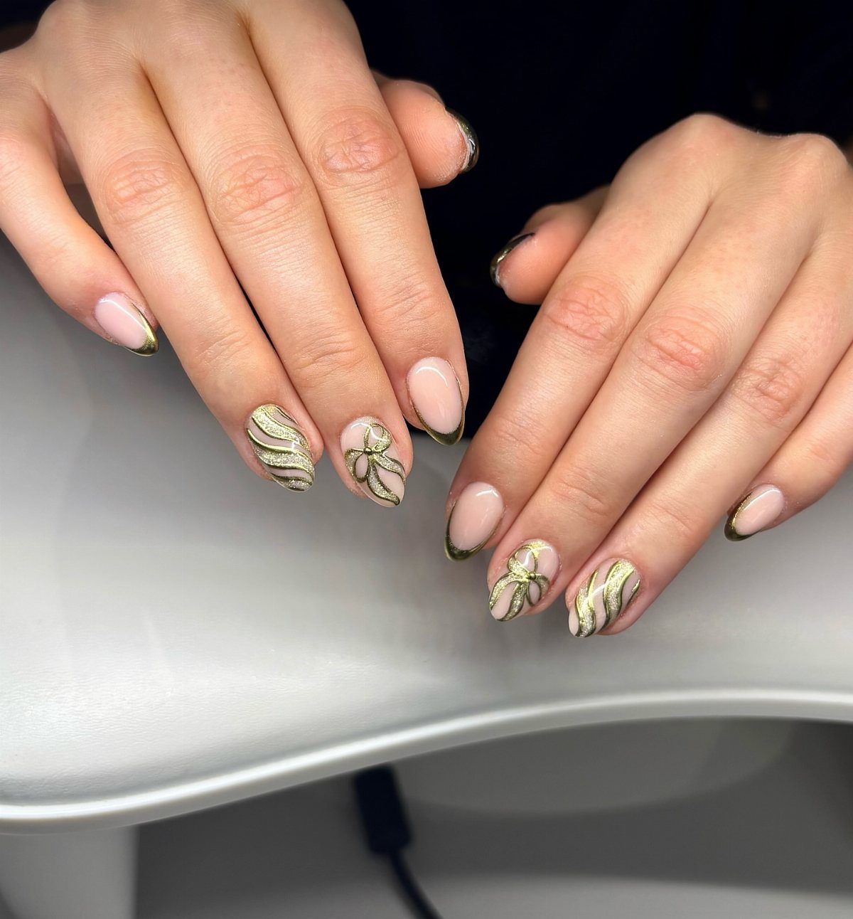 Nude almond nails with gold glitter French tips and decorative glitter bow designs.
