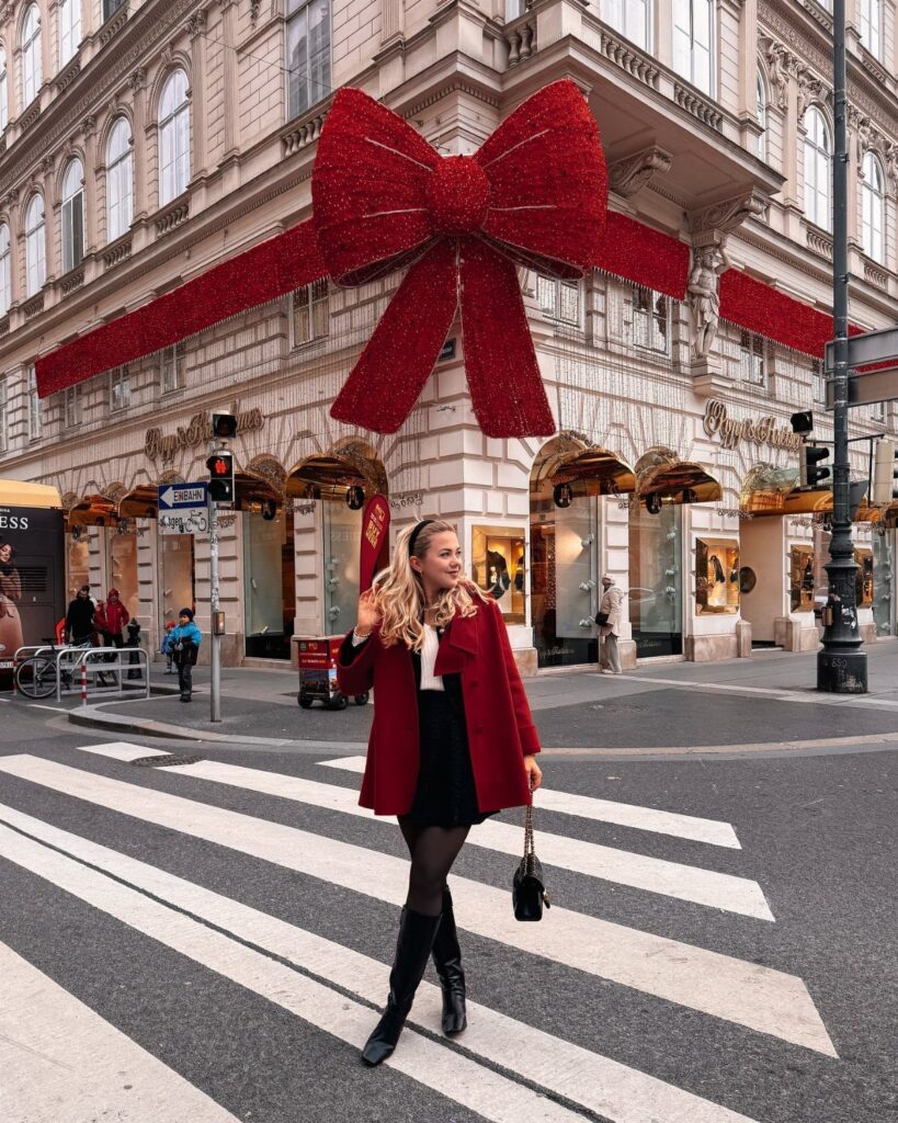 Woman in a red coat and black skirt standing before a giant red bow.