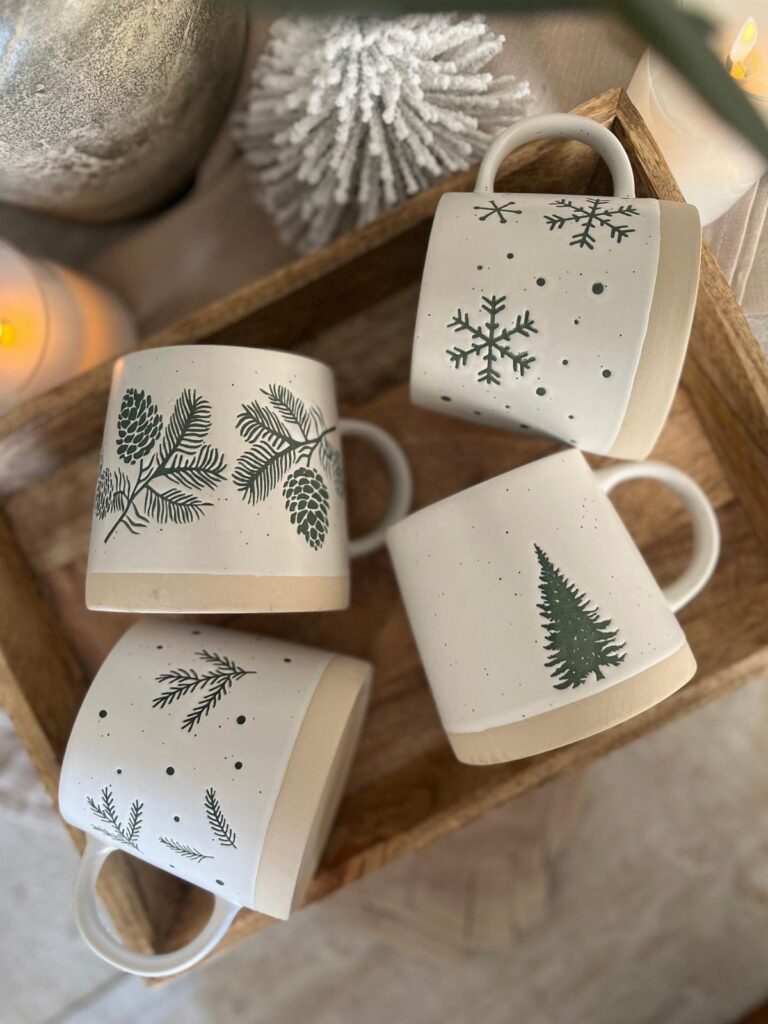 Four cream ceramic mugs with green winter designs sitting in a wooden tray.