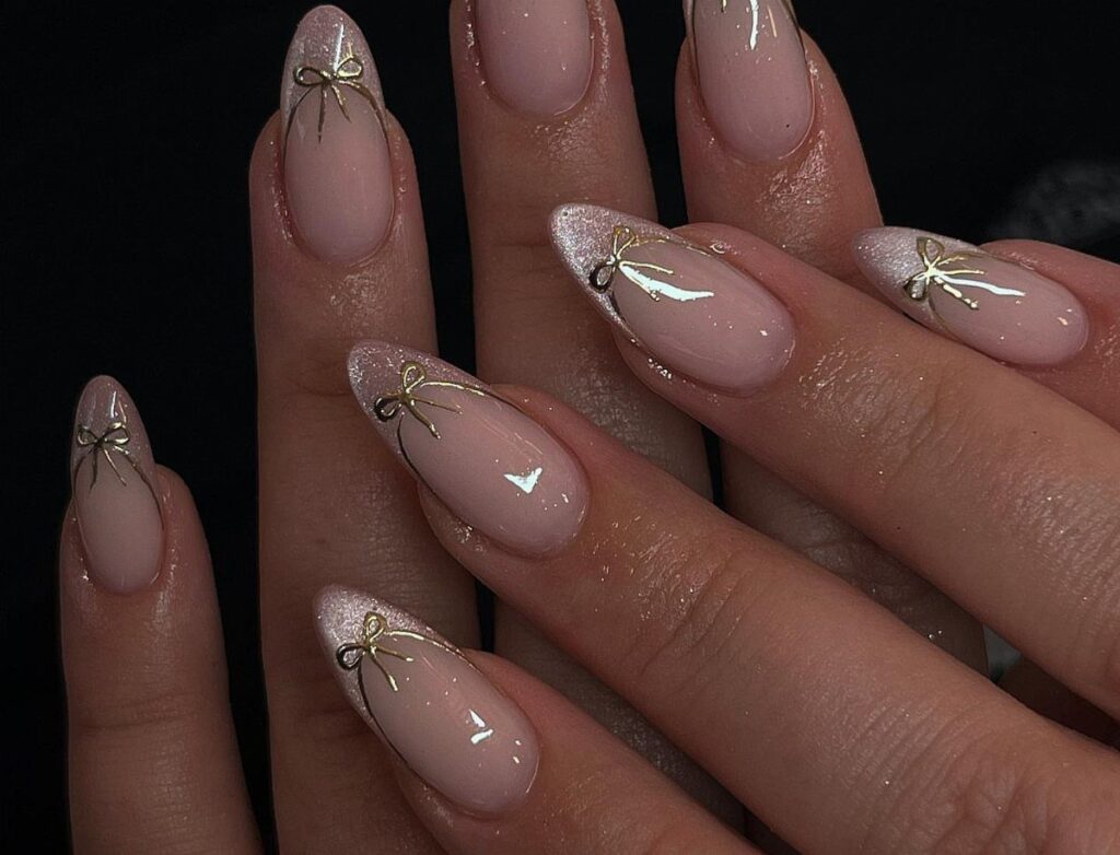 Nude almond nails with silver glitter tips and gold chrome bow accents.