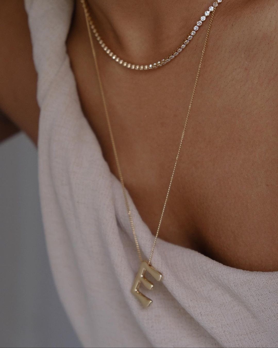 Gold initial necklace with letter E and a diamond tennis necklace on a woman