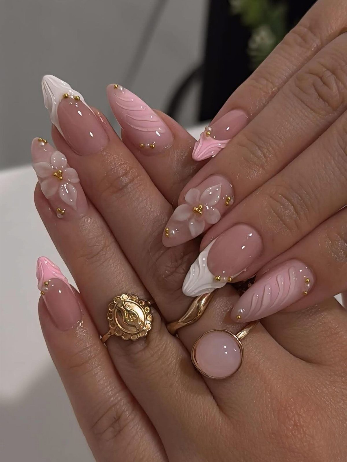 Almond-shaped nails with pink polish, 3D white flowers, and gold accents on hands.