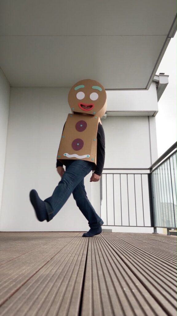 Person in a homemade brown cardboard gingerbread man costume walking on a wooden deck
