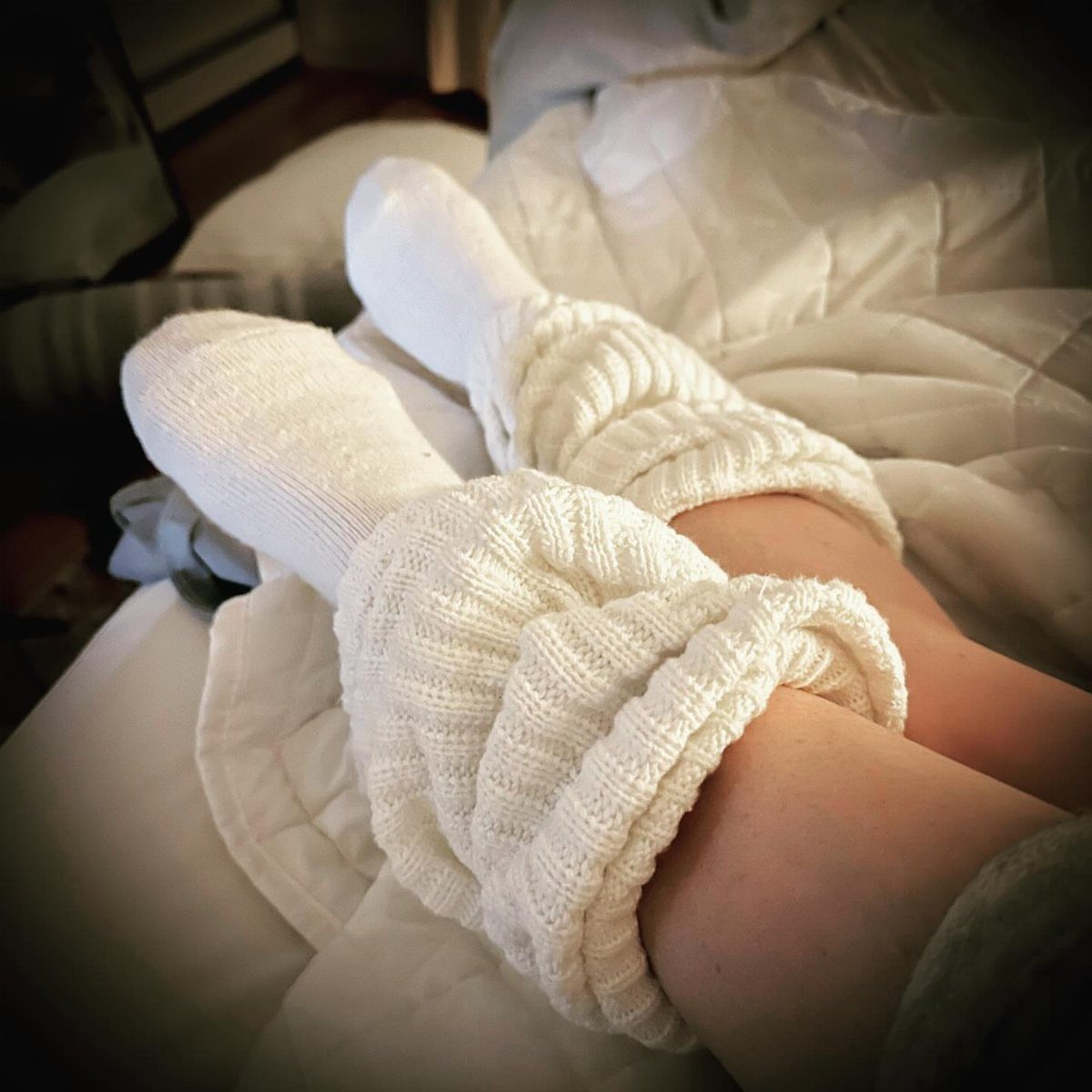 White chunky knit cozy socks worn on feet resting on a white bed.