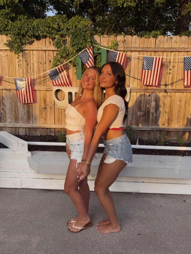 Two women in white crop tops and denim shorts posing by American flags.
