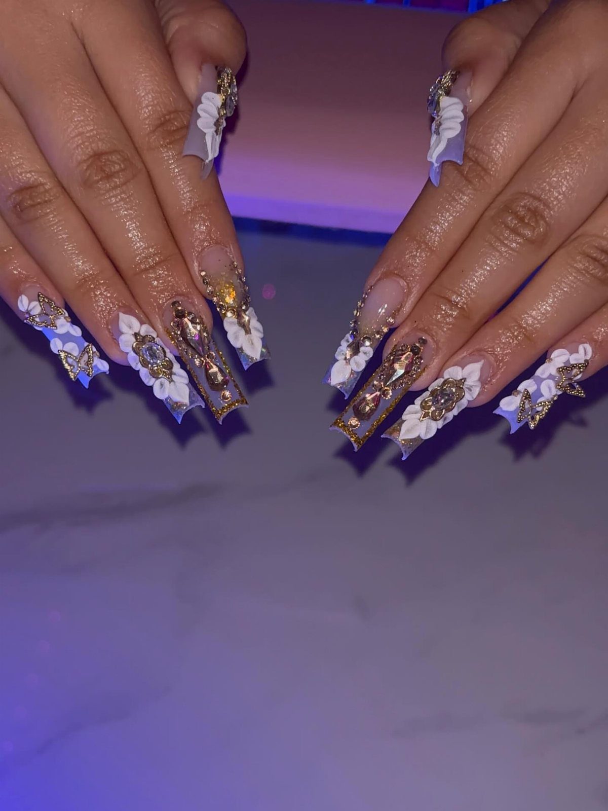 Long acrylic nails with white 3D flowers and ornate gold glitter accents.