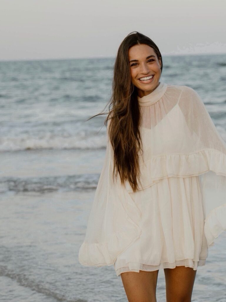 Woman in a white flowy tiered dress smiling on a sandy beach shoreline.