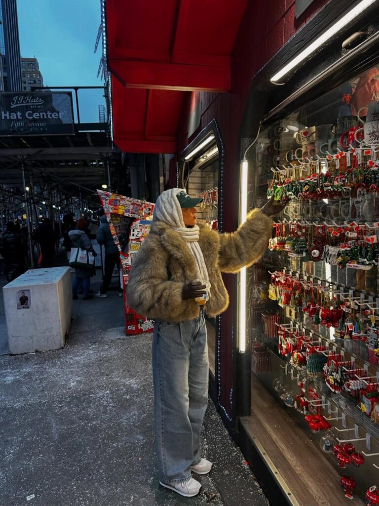 Person in faux-fur jacket and jeans browsing Christmas ornaments in a city window.