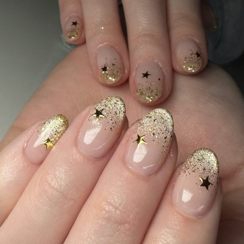 Nude nails with gold glitter ombre tips and small gold star accents.