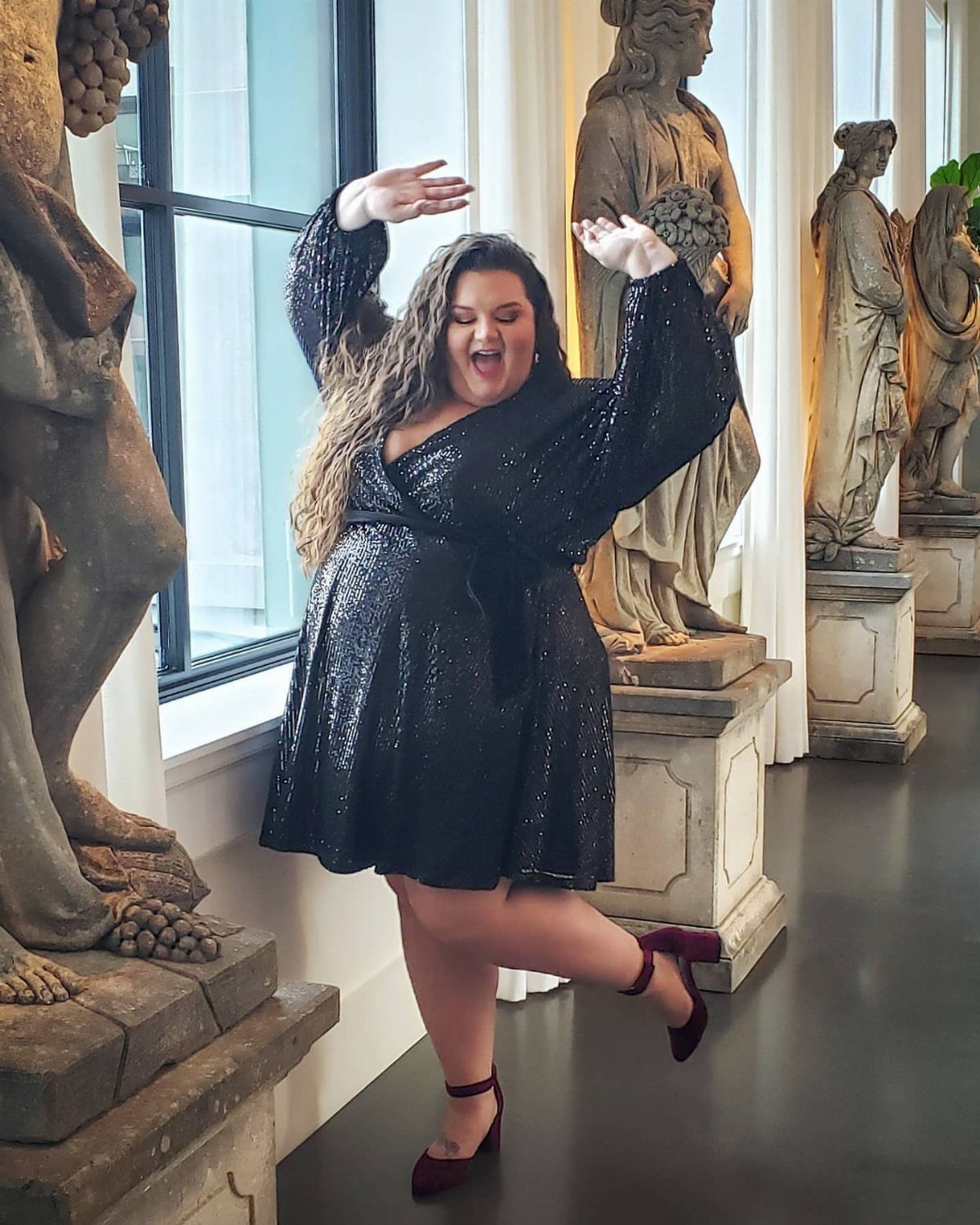 Woman in a black sequin dress posing in a gallery with stone statues.