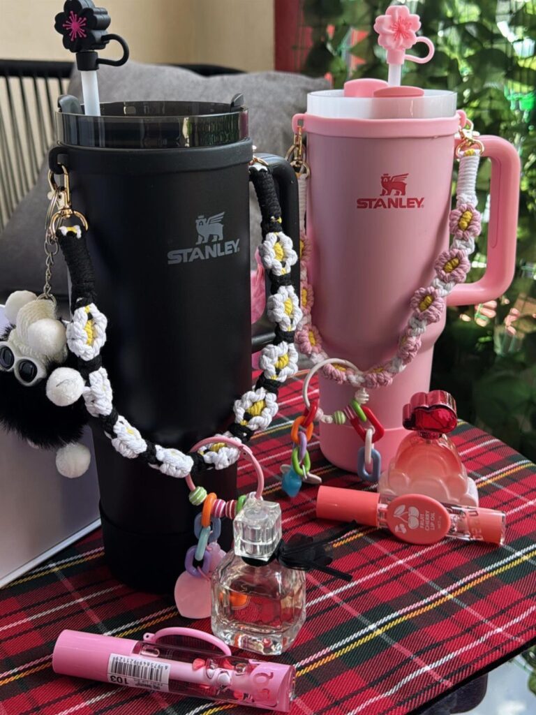Black and pink Stanley water bottles with crocheted flower straps on plaid fabric.