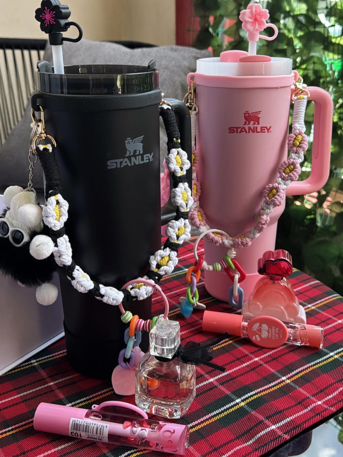 Black and pink Stanley water bottles with crocheted flower straps on plaid fabric.