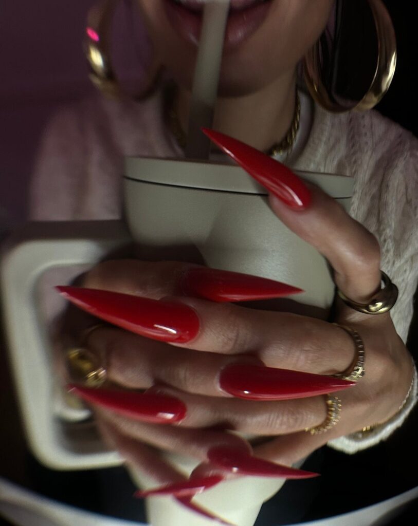 Long red stiletto nails holding a beige cup with gold jewelry accents.
