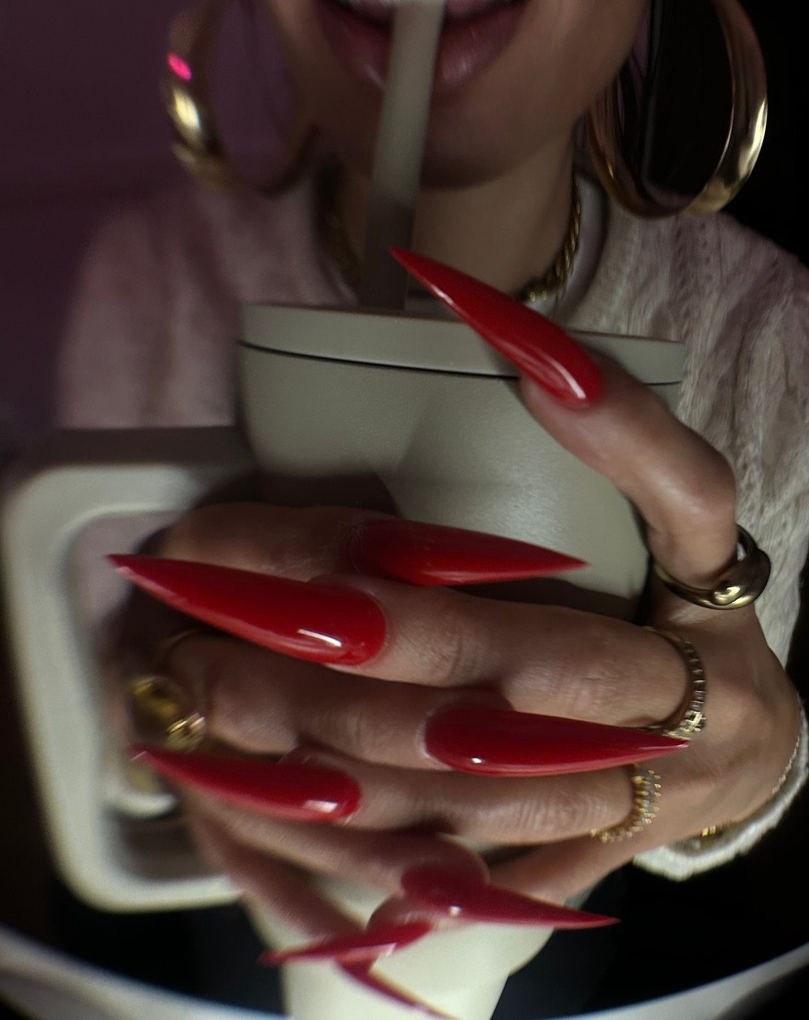 Long red stiletto nails holding a beige cup with gold jewelry accents.