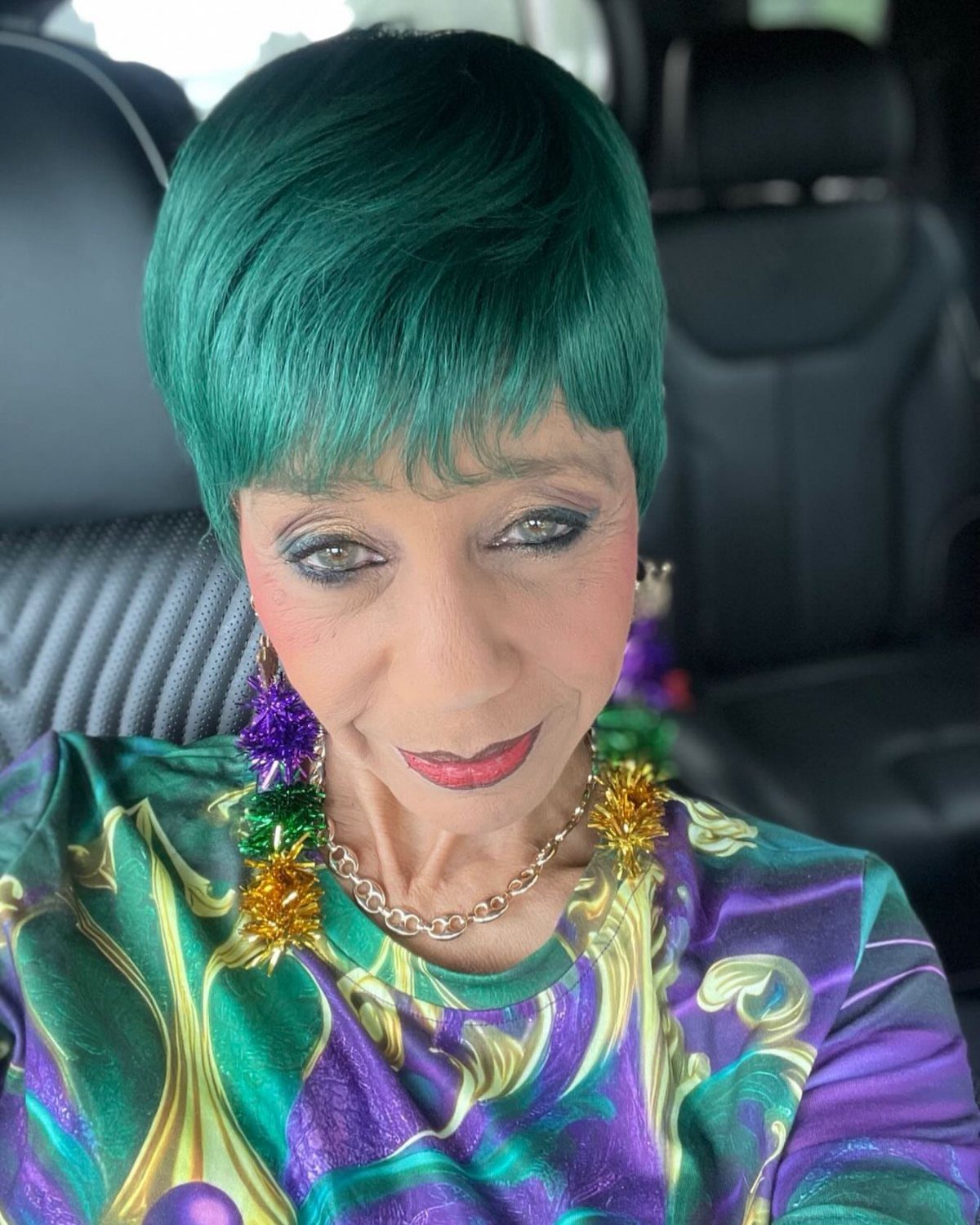Woman with green hair wearing a colorful Mardi Gras themed outfit and earrings.